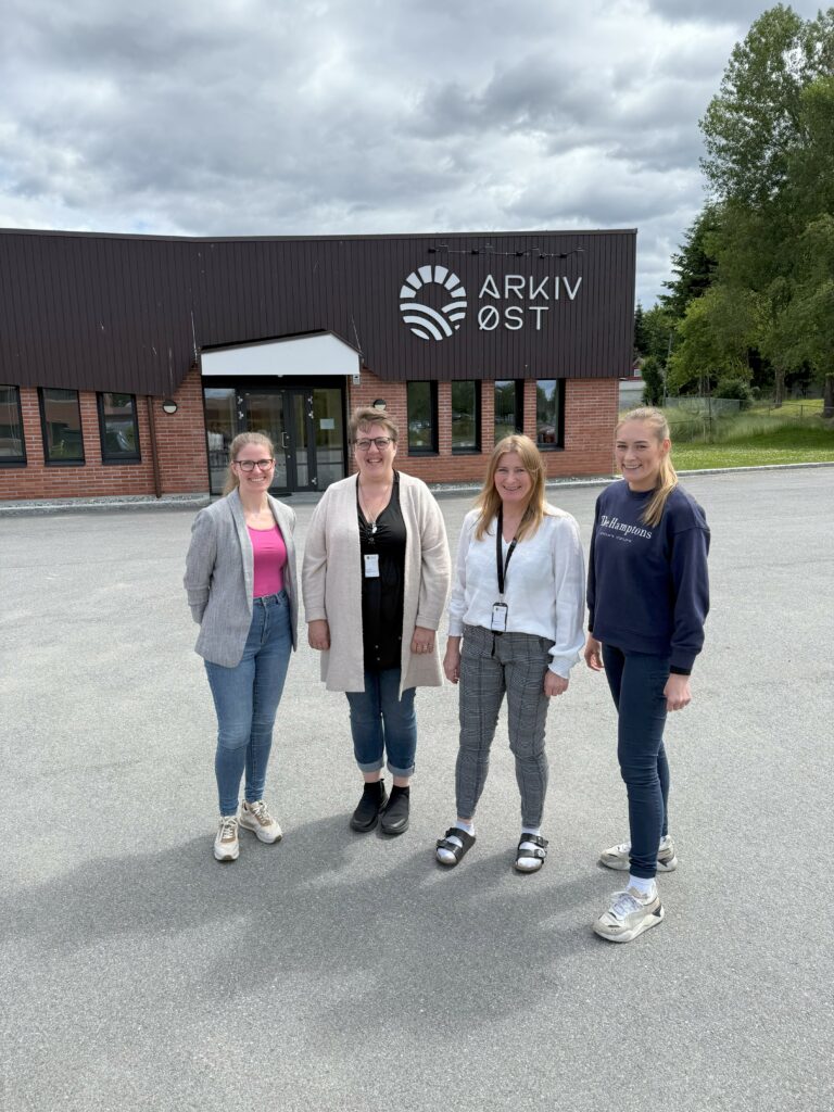 Bildet viser fire damer som står foran et murbygg. På veggen synes logoen til Arkiv Øst.